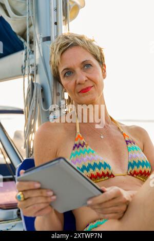 Eine Frau mit kurzen blonden Haaren, die ein farbenfrohes Bikinioberteil trägt, hält eine Tablette, während sie auf einem Segelboot mit nachdenklichem Ausdruck sitzt. Stockfoto