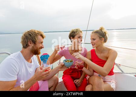 Drei Freunde genießen Getränke und Snacks auf einem Boot an einem sonnigen Tag mit klarem Himmel und einem ruhigen See im Hintergrund. Stockfoto