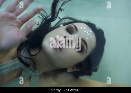 Eine junge Frau mit langen, dunklen Haaren schwimmt anmutig in einem Aquarium, ihre Augen geschlossen, als wären sie in einem Traum verloren. Stockfoto