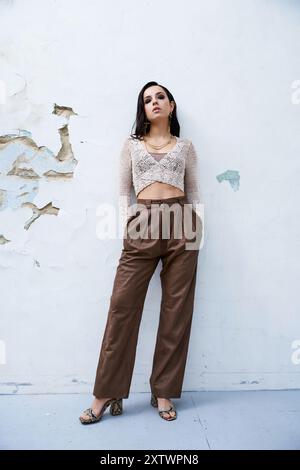 Eine junge Frau mit langen brünetten Haaren posiert in einem weißen Strickoberteil und einer braunen Hose vor einer weißen Wand. Stockfoto