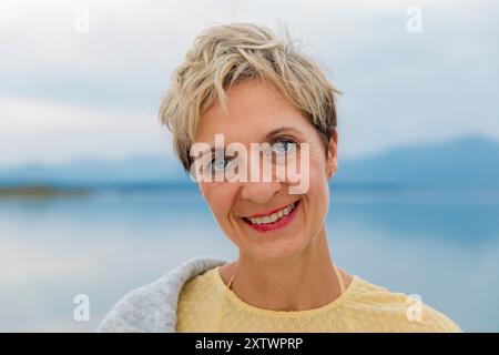 Lächelnde Frau mit kurzen blonden Haaren draußen vor einem verschwommenen Hintergrund am Wasser. Stockfoto