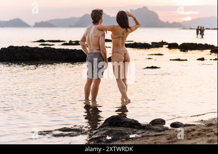 Ein Paar hält die Hände, während er bei Sonnenuntergang an einem Strand spaziert, wobei das Meer sanft zu ihren Füßen und Berge in der Ferne liegen. Stockfoto