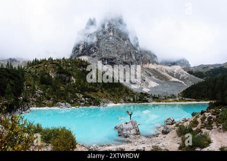 Eine Person steht auf einem Felsen mit triumphalen Armen vor einem atemberaubenden türkisfarbenen Bergsee mit einem majestätischen Berg im Hintergrund. Stockfoto