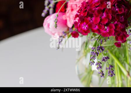 Leuchtender Blumenstrauß aus rosa Pfingstrosen und lila Blumen in einer klaren Vase auf einer weißen Oberfläche mit verschwommenem Hintergrund. Stockfoto