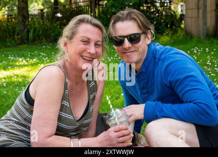 Lächelnde Frau und Mann sitzen auf dem Gras und genießen einen sonnigen Tag im Freien, während der Mann einen Drink hält. Stockfoto