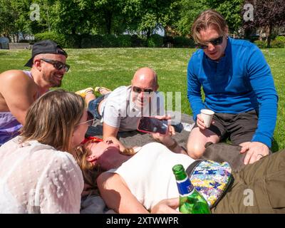 Freunde lachen zusammen, während sie auf einem grasbewachsenen Park liegen, während einer etwas auf einem Smartphone zeigt. Stockfoto