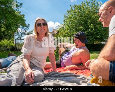 Lächelnde Frau in Sonnenbrille, die einen sonnigen Tag draußen bei einem Picknick mit zwei Männern genießt. Stockfoto