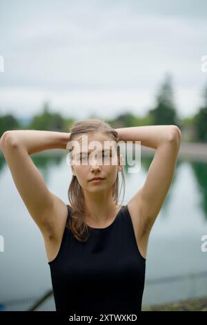 Die junge Frau in einem schwarzen Dach steht selbstbewusst mit den Händen hinter dem Kopf vor einem verschwommenen See-Hintergrund. Stockfoto