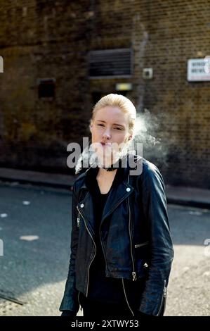 Frau, die Rauch ausatmet, während sie im Sonnenlicht gegen eine Ziegelwand steht und eine schwarze Lederjacke trägt. Stockfoto