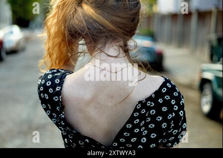 Eine Frau mit lockigen roten Haaren, die ein schwarzes Kleid mit weißen Blumen trägt, steht mit dem Rücken zur Kamera auf einer Stadtstraße. Stockfoto