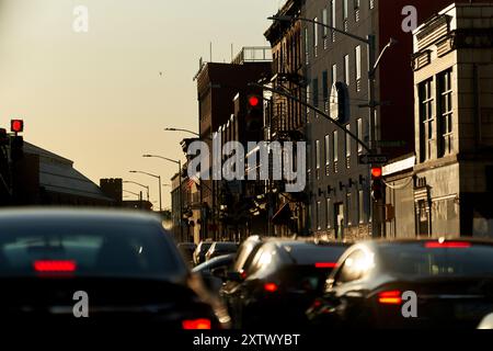 Abendlicher Verkehr auf einer Stadtstraße mit roten Ampeln und der Sonnenuntergang hinter Gebäuden, der ein warmes Leuchten ausstrahlt. Stockfoto