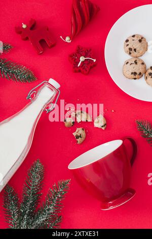 Trendiges Weihnachtslayout mit Kaffeetasse, Milchflasche, Schokoladenkeksen, Weihnachtsbaumzweigen und roten Weihnachtskerzen auf rotem Hintergrund. Stockfoto