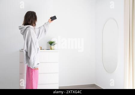 Eine junge Frau hebt den Arm, um ein Selfie zu machen. Sie ist in ihrem Zimmer zu Hause und trägt ein graues Sweatshirt und eine pinkfarbene Hose. Sie ist vor einem M Stockfoto