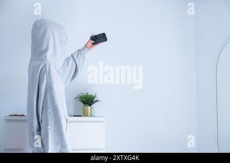 Eine junge Frau hebt den Arm, um ein Selfie zu machen. Sie ist in ihrem Zimmer zu Hause und trägt ein graues Sweatshirt und eine pinkfarbene Hose. Sie bedeckt ihr Gesicht mit Stockfoto