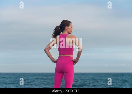 Eine Frau mit Händen auf den Hüften und Blick auf das Meer in rosafarbener Sportbekleidung. Entspannung nach dem Training. Stockfoto