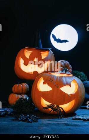 Zwei beleuchtete Jack-o'-Laternen mit gruseligen Ausdrücken, eine mit Hexenhut, unter Vollmond mit Fledermaus-Silhouette Stockfoto