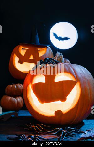 Zwei leuchtende Jack-O'-Laternen, gruselige Kürbis-Gesichter, Vollmond und Fledermaus im Hintergrund schaffen eine gruselige Halloween-Szene Stockfoto