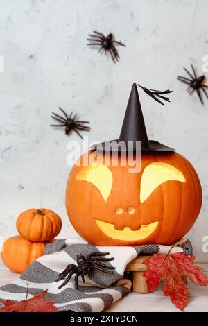 Glühend geschnitzter Kürbis mit freundlich geschnitztem Gesicht im Hexenhut, Herbstblättern, Minikürbissen und Spinnen Stockfoto