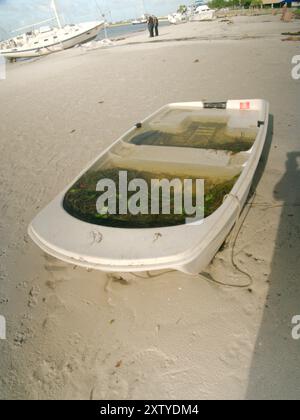 Kleines weißes Ruderboot gefüllt mit Wasser, das nach einem großen Sturm am Ufer lag. Gulfport Florida mit dem Strand und dem blauen Himmel im Hintergrund. Ein Stockfoto