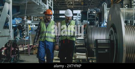 Zwei professionelle Schwerindustrietechniker in Schutzuniform und Helmen diskutieren über den Herstellungsprozess in der Fabrik. Männliche Mitarbeiter arbeiten im Werk. Zeitlupe. Stockfoto