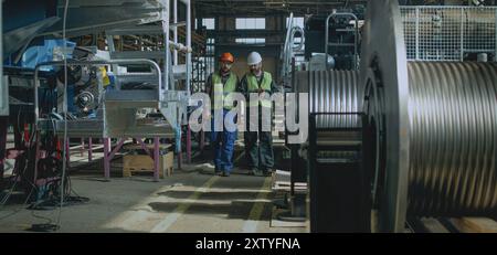 Zwei professionelle Schwerindustrietechniker in Schutzuniform und Helmen diskutieren über den Herstellungsprozess in der Fabrik. Männliche Mitarbeiter arbeiten im Werk. Zeitlupe. Stockfoto