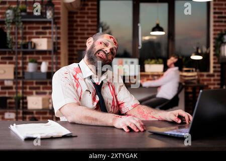 Erschöpfter Mann sackte über den Schreibtisch, umgeben von Papierkram, wurde zu Tode bearbeitet. Mitarbeiter im Amt wurden zu Zombie, Konzeptualisierung des unerbittlichen Drucks kapitalistischer Jobs Stockfoto