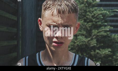 Filmische Aufnahme eines ernsthaften Teenagers, der in die Kamera schaut, während er draußen bei starkem Regen steht. Regentropfen laufen dem Teenager ins Gesicht. Nadelbaum im Hintergrund. Hochformat. Zeitlupe. Stockfoto