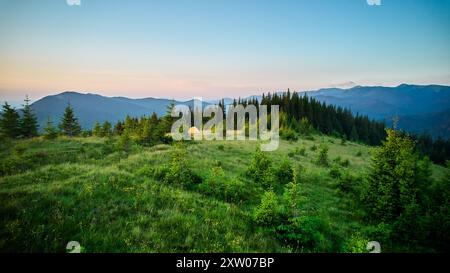 Hellgelbes Touristenzelt auf einem grasbewachsenen Hügel, umgeben von üppigen grünen Kiefern. Atemberaubender Blick auf die fernen blauen Berge unter klarem Himmel, die einen idyllischen und ruhigen Campingplatz schaffen. Stockfoto