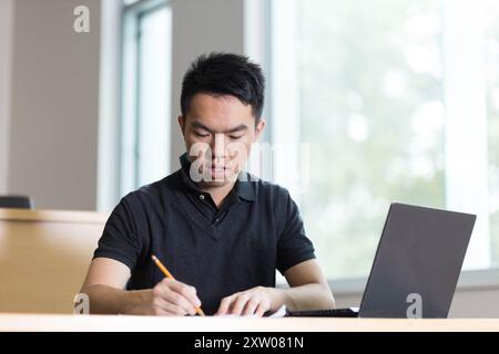 Ein junger Student sitzt gerade an einem Tisch und schreibt mit seinem Bleistift auf ein Blatt Papier mit seinem Laptop neben ihm Stockfoto