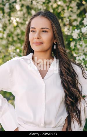 Porträt eines schönen Mädchens mit langen Haaren in der Nähe eines blühenden Baumes in weißen, trendigen Kleidern Stockfoto