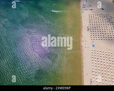 Drohnenvideo, das einen Blick von oben auf einen Strand mit Reihen von bunten Sonnenschirmen aufnimmt, die perfekt auf dem Sand neben der Küste angeordnet sind. Der lebendige Stockfoto