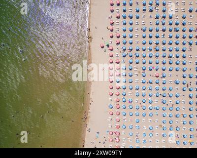 Drohnenvideo, das einen Blick von oben auf einen Strand mit Reihen von bunten Sonnenschirmen aufnimmt, die perfekt auf dem Sand neben der Küste angeordnet sind. Der lebendige Stockfoto