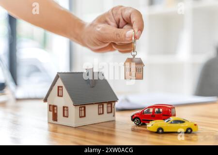 Nahhand eines Mannes, der einen Hausschlüsselanhänger mit einem Haus hält und Autos Modelle auf einem Schreibtisch hat, Haus oder Immobilien kauft Stockfoto