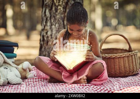 Magie, Mädchen und Lesebuch über Picknick im Wald für Geschichte, Fantasie und Roman für die kindliche Entwicklung. Weibliche Person, draußen und lächeln mit Spielzeug Stockfoto