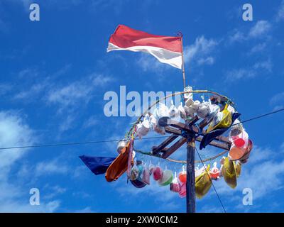 Verschiedene Preise hängen an der Spitze einer Stange, die für einen Panjat pinang Wettbewerb verwendet wird. Über ihnen flattert die rot-weiße Fahne gegen einen hellblauen Himmel. Stockfoto