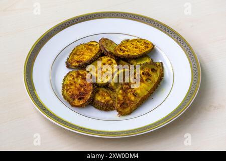 Gebratener Kürbis oder Kakrol bhaja auf einer weißen Platte auf hellem Holz. Momordica dioica, auch bekannt als Kürbis, Käntola, Kartoli, Bhat Karela, Stockfoto