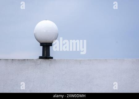 Weiße runde Glaslampe auf einem schwarzen Stahlmast an der Hauswand, heller himmelblauer Hintergrund. Stockfoto