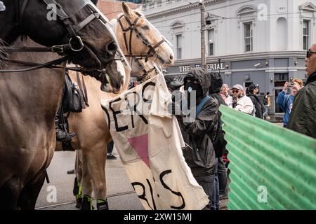 Melbourne, Australien. August 2024. Ein maskierter Demonstrant hält ein Banner vor den berittenen Polizeibeamten während einer Demonstration treffen sich rechts-Trans-Aktivisten mit der Polizei, als sie eine Anti-Trans-Gruppe im Parlamentsgebäude in melbourne konterkarieren, Verhaftungen durchgeführt, alberne Schnur und Wasserblasen von den Gegenprotestierenden auf Polizeibeamte verwendet wurden. Quelle: SOPA Images Limited/Alamy Live News Stockfoto