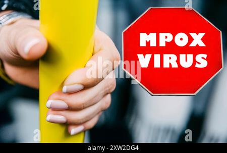 Eine Hand hält einen gelben Stab, während ein rotes Warnschild über den Mpockenvirus in der Nähe in einer öffentlichen Verkehrsmittel-Umgebung deutlich sichtbar angezeigt wird. Stockfoto