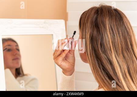 Eine junge blonde Frau, die vor einem Spiegel steht, trägt Mascara auf die Wimpern. Das Bild fängt einen fokussierten Moment ihres M ein Stockfoto