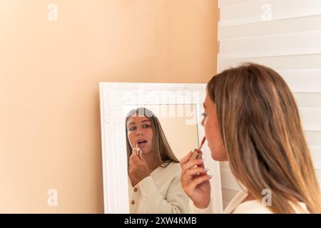 Die junge blonde Frau, die vor einem Spiegel steht, trägt Lippenstift auf. Das Bild fängt einen wichtigen Moment in ihrer Make-up-Routine, Focus, ein Stockfoto