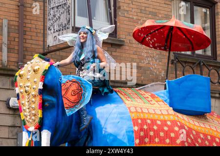 Belfast Mela Carnival Parade - glückliche Frau mit blauen Haaren und Feenflügeln, die auf blauem Elefanten mit goldener Kopfdekoration reitet. Belfast, Großbritannien - 17. August 2024. Stockfoto