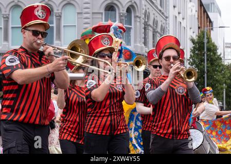 Belfast Mela Carnival Parade – Samba-Band in unverwechselbaren roten und schwarzen Hemden unterhält Menschenmassen im Stadtzentrum. Belfast, Großbritannien - 17. August 2024. Stockfoto