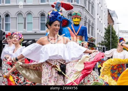 Belfast Mela Karnevalsparade: Frauen in festlichen Kostümen mit dekorativen Blumen im Haar tanzen durch das Stadtzentrum. Belfast, Großbritannien - 17. August 2024. Stockfoto