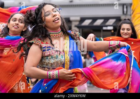 Belfast Mela Carnival Parade: Drei junge indische Frauen in farbenfroher traditioneller Kleidung tanzen im Stadtzentrum. Belfast, Großbritannien - 17. August 2024. Stockfoto