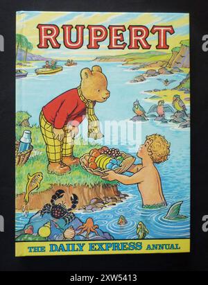 Ein Jahrgang „Rupert“ für Kinder, der 1975 von der Tageszeitung Daily Express veröffentlicht wurde. Das jährliche Jahr basiert auf der beliebten englischen Comicfigur Rupert Bear, die 1920 erstmals im Daily Express erschien. Das farbenfrohe Cover zeigt Rupert am Meer und nimmt eine große Schale mit Früchten eines Meerjungfrauenjungen auf. Stockfoto