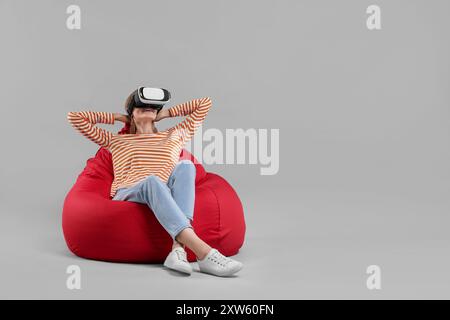 Glückliche Frau mit Virtual-Reality-Headset auf Sitzsack-Stuhl vor grauem Hintergrund, Platz für Text Stockfoto