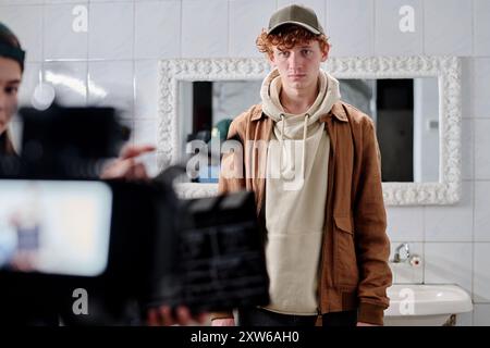 Porträt eines jungen Mannes, der vor dem Badezimmerspiegel mit lockigem Haar steht und Hoodie und Jacke trägt, während ein Mitglied der Filmcrew die Kamera im Vordergrund hält Stockfoto