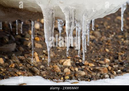 Eiszapfen hängen von einer Eisscholle am Strand, die über einem Kiesstrand hängt. Am Strand, im Vordergrund liegt Schnee. Stockfoto