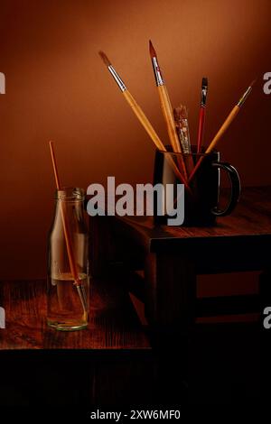 Bürsten in Gouache und Flasche Wasser auf Holztisch vor warmem rustikalem Hintergrund Stockfoto